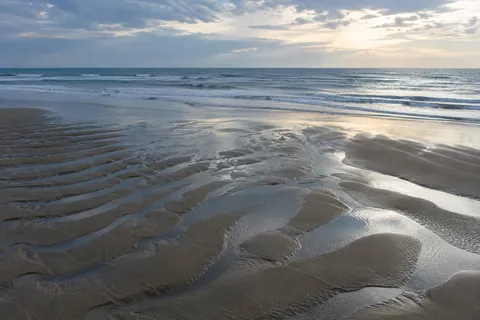 Shallow reflective channels wind among broad low-tide sand ridges beneath a clouded coastal sky.