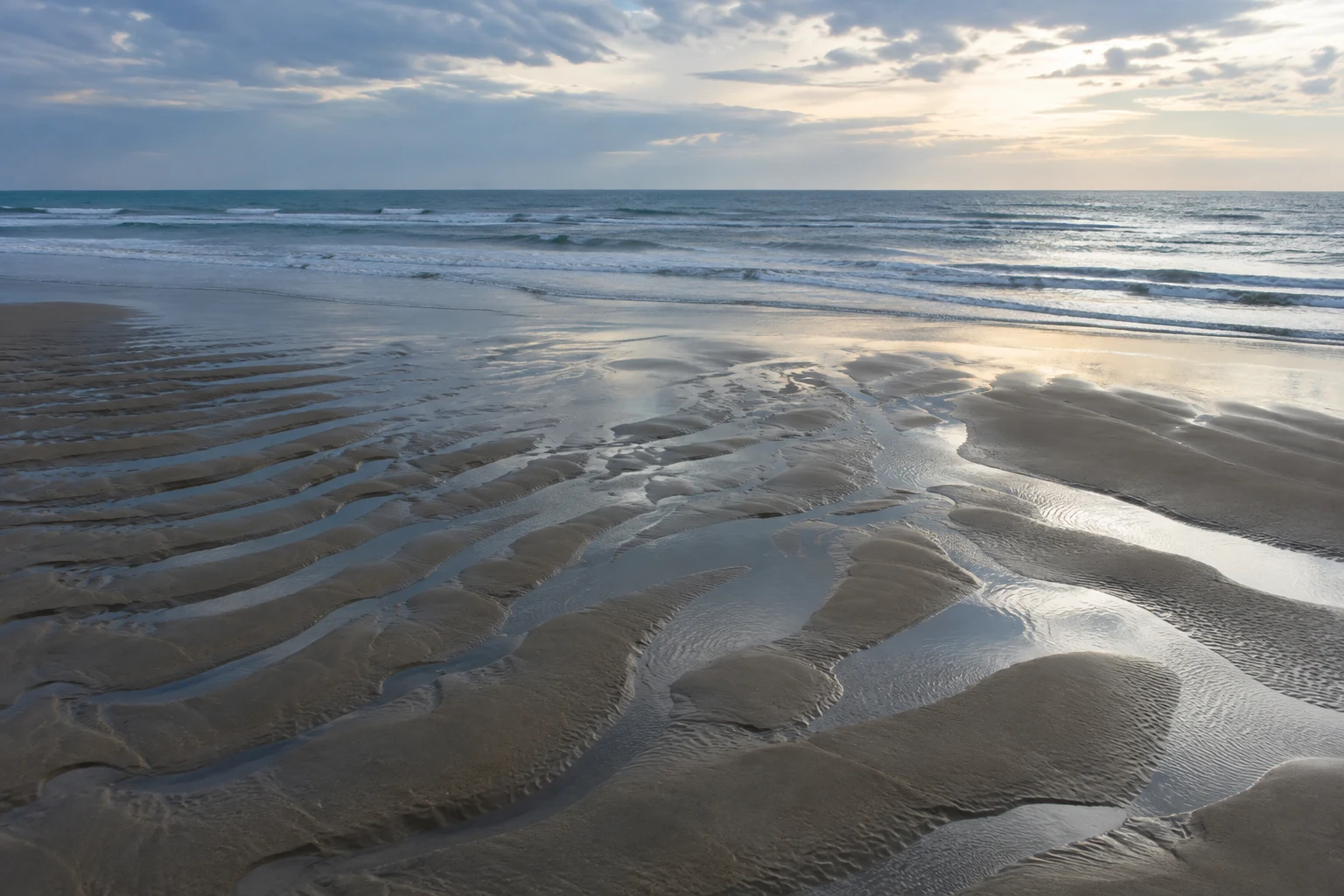 Shallow reflective channels wind among broad low-tide sand ridges beneath a clouded coastal sky.