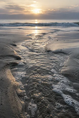A narrow silver-gold channel runs through wet sand toward distant surf and a low warm sun.