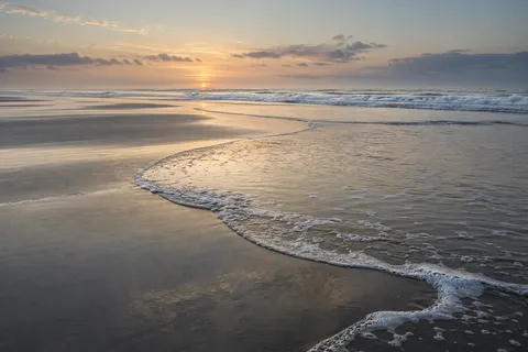A sweeping curve of shallow backwash reflects pale evening light across an empty beach.