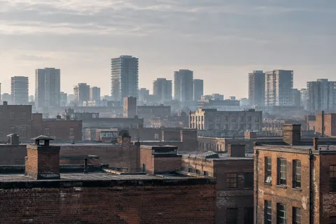 Weathered brick rooftops and distinct chimney stacks below a hazy skyline of gray towers.