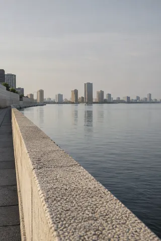 A textured pale river wall recedes diagonally beside calm water toward anonymous towers.