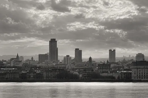 A monochrome skyline of unequal towers and older stone buildings beneath dramatic clouds beside a bright river.