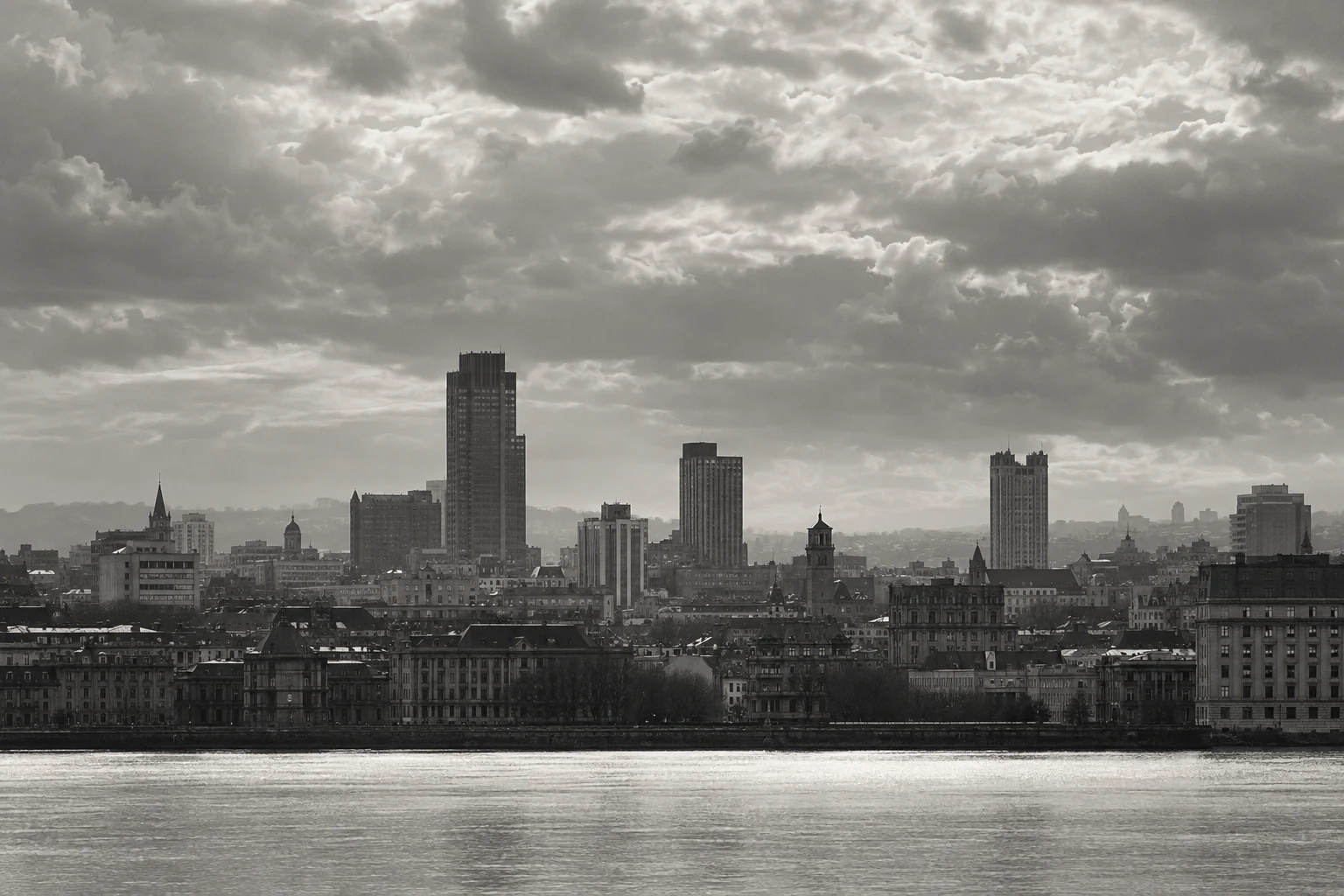 A monochrome skyline of unequal towers and older stone buildings beneath dramatic clouds beside a bright river.