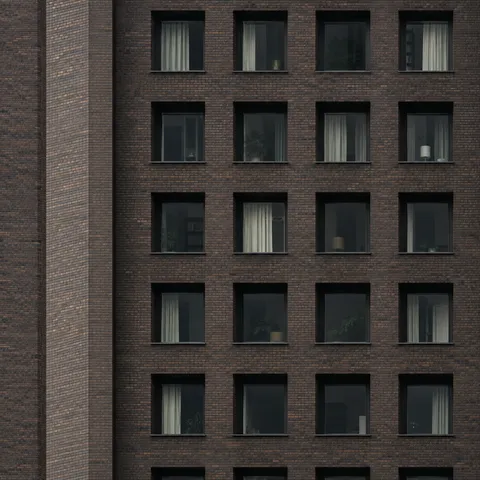 A close view of dark brown brickwork with aligned square windows and varied pale curtains.
