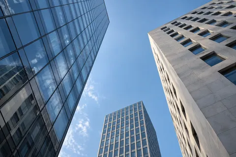 An upward view between a reflective glass tower, pale stone facade and smaller distant tower.