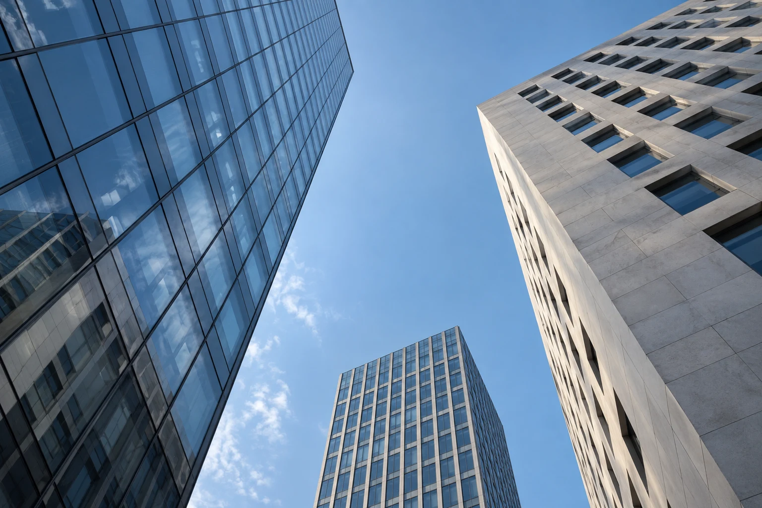An upward view between a reflective glass tower, pale stone facade and smaller distant tower.
