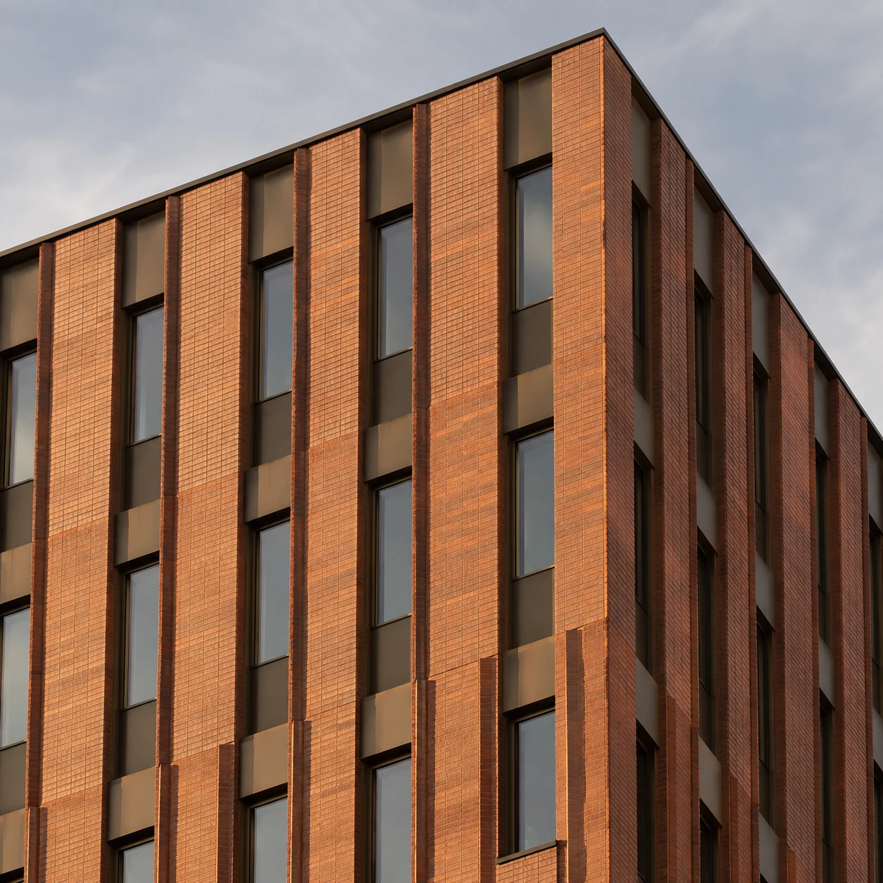 An oblique close view of a terracotta brick corner with tall window bays against soft gray sky.