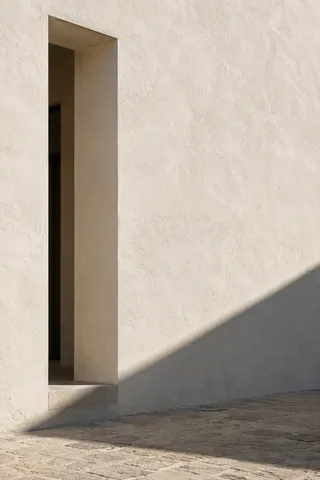A tall recessed opening and diagonal shade on a warm white plaster wall above worn paving.