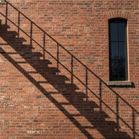 A stair and railing shadow on an old brick facade beside one narrow dark window.