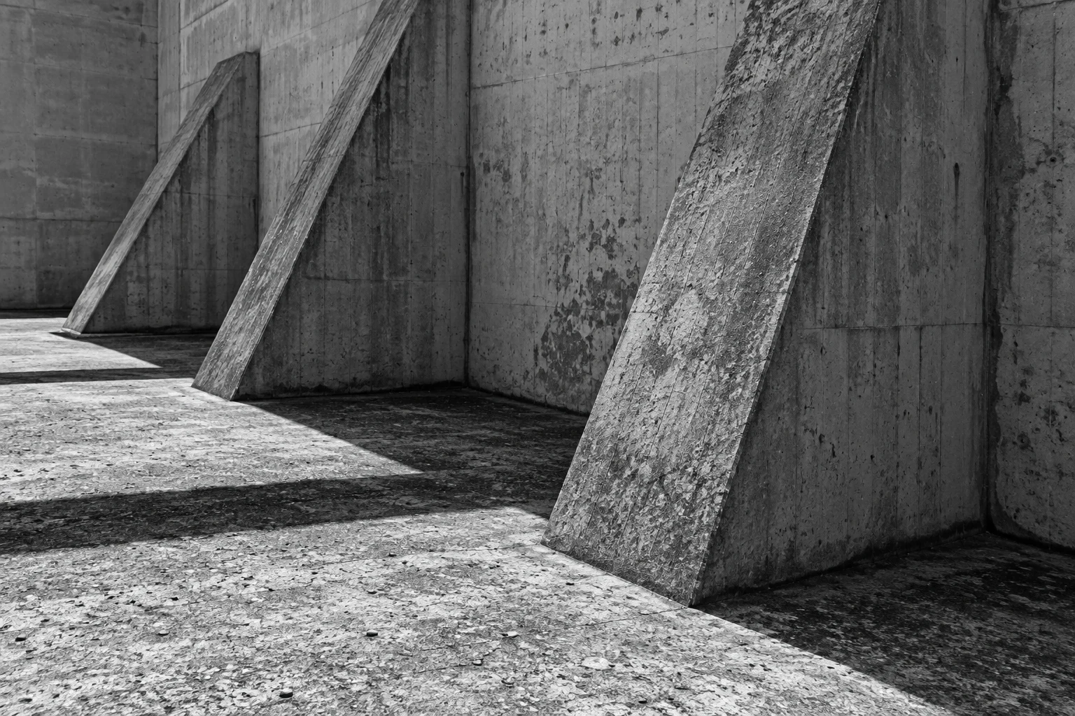 A monochrome close view of three sloping concrete buttresses and long shadows.