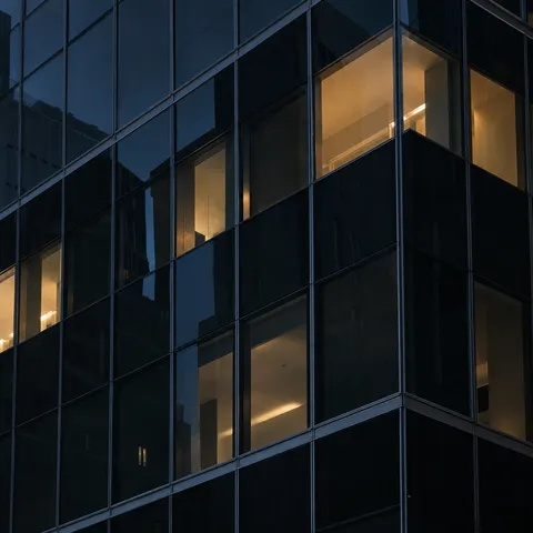 An oblique nighttime glass corner with scattered amber-lit empty interiors and subtle neighboring reflections.