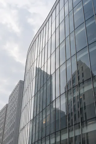 An upward view of a curved silver-blue glass building reflecting anonymous pale towers.