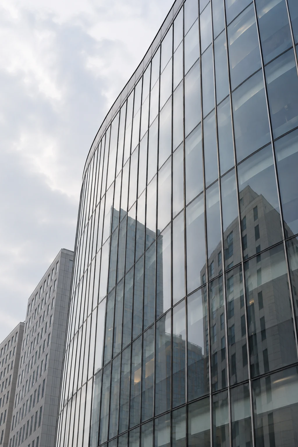 An upward view of a curved silver-blue glass building reflecting anonymous pale towers.