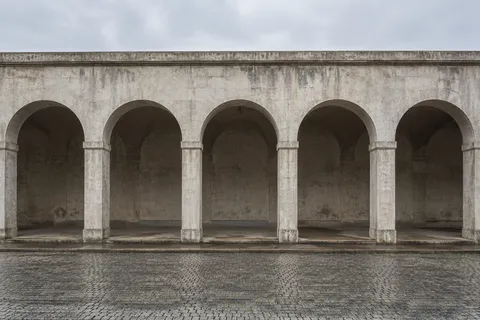 A frontal view of an empty weathered arcade with five broad arches and dark recessed bays.