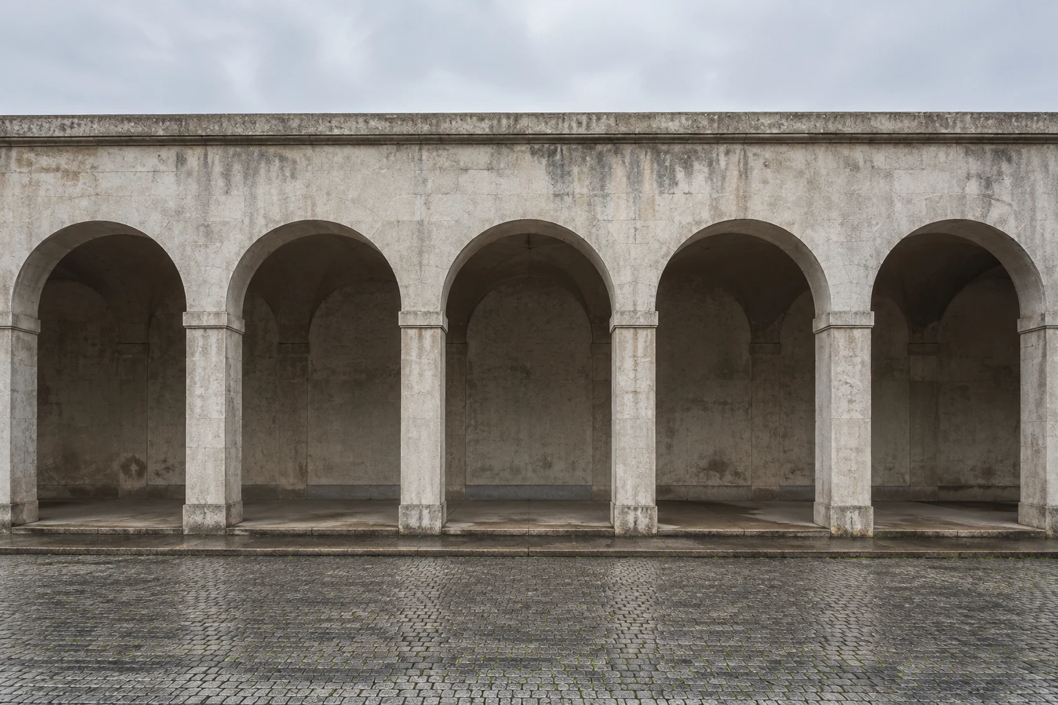 A frontal view of an empty weathered arcade with five broad arches and dark recessed bays.