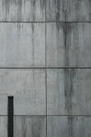 A close concrete panel facade with fine joints, uneven rain stains and a narrow dark ventilation grille.