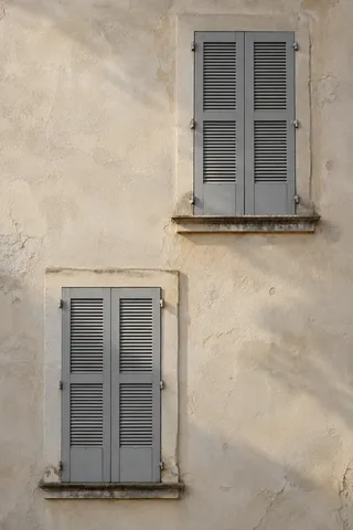 Two closed gray shuttered windows placed diagonally on aged cream plaster with stone sills.