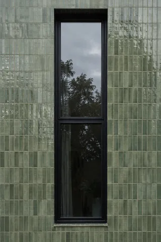 One tall black window in a muted green tiled facade with tree and cloud reflections.