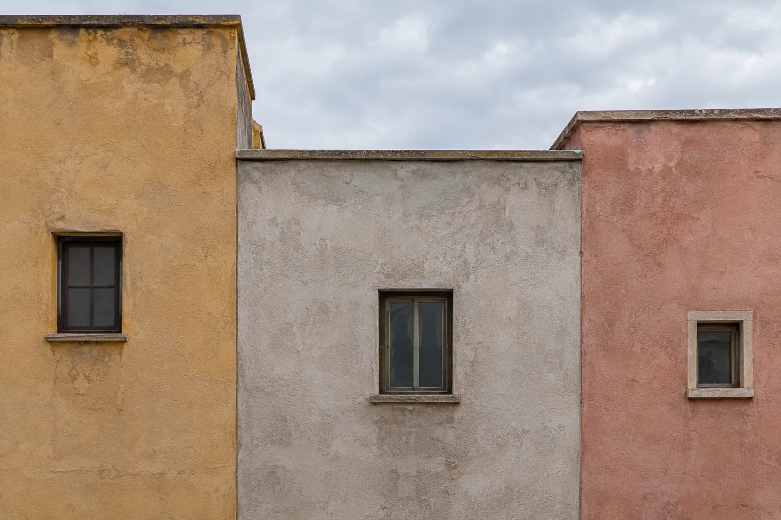 Three adjoining muted stucco facades with different roof heights and small dark windows beneath cloudy sky.