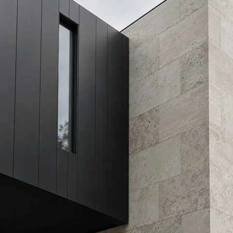 A charcoal metal facade with a slender window beside pale limestone blocks, seen at an upward angle.