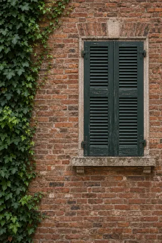 A closed dark green shuttered window in warm brick with an irregular column of ivy along the left edge.