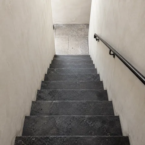 A downward view of charcoal stone stairs between cream walls with a dark handrail on the right.