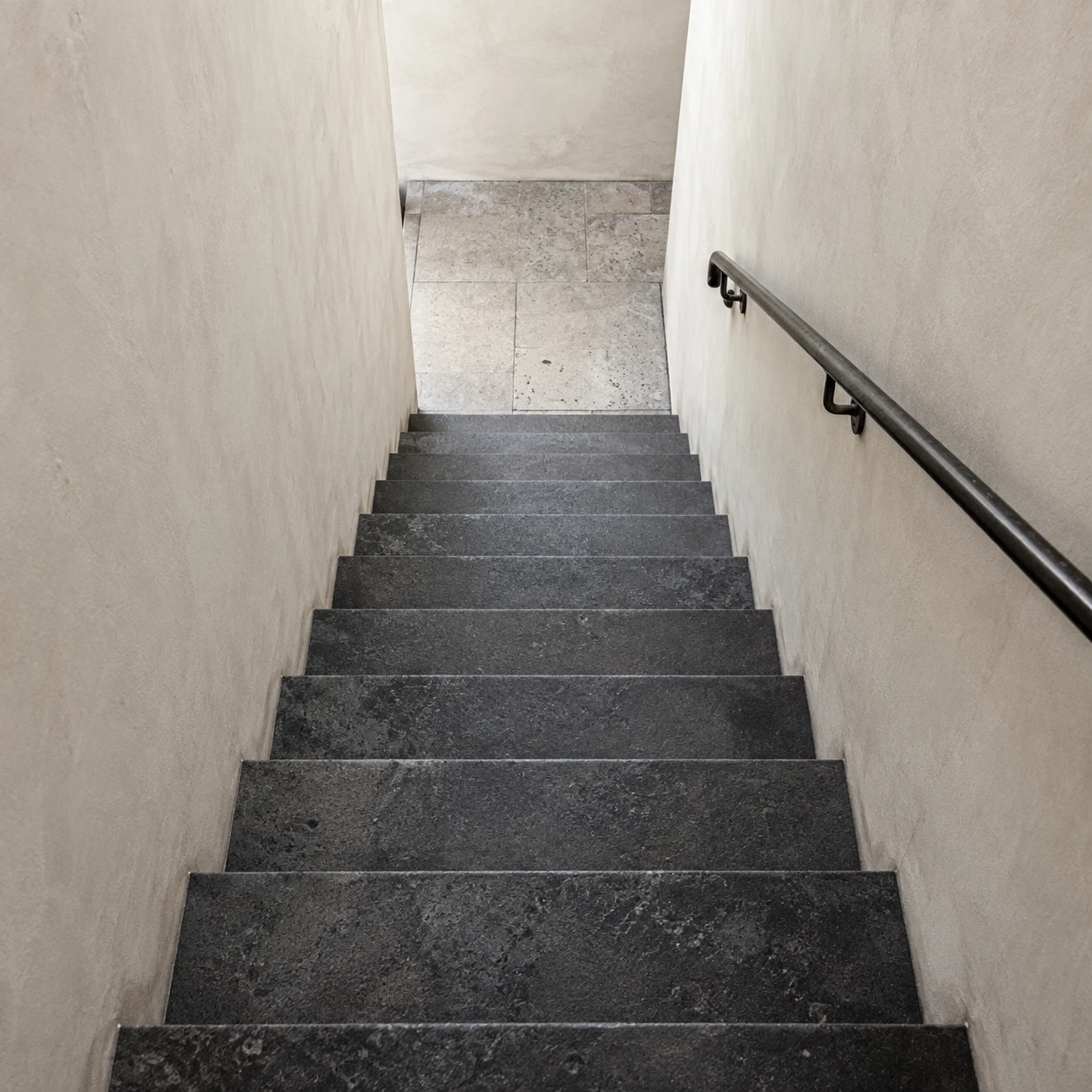 A downward view of charcoal stone stairs between cream walls with a dark handrail on the right.