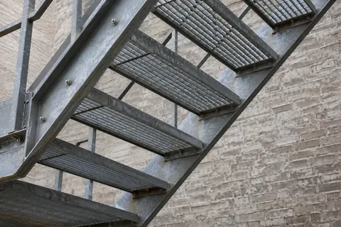 A close view from beneath a steel stair with open grating treads, bolted stringers and pale brick behind.