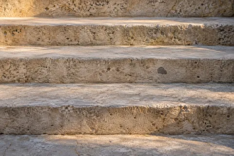 A close crop of aged limestone stair treads with rounded edges, fine pits and warm patches of sunlight.
