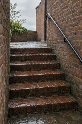 Wet red-brick steps rise between brick walls to an empty landing, with a black handrail on the right.