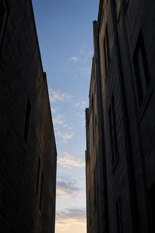 An upward view between two tall shadowed masonry buildings framing a narrow blue sky with pale clouds.