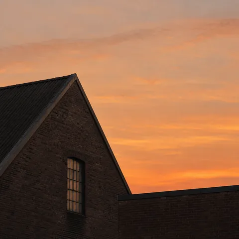 A steep warehouse gable and lower flat brick roof against softly glowing apricot clouds.