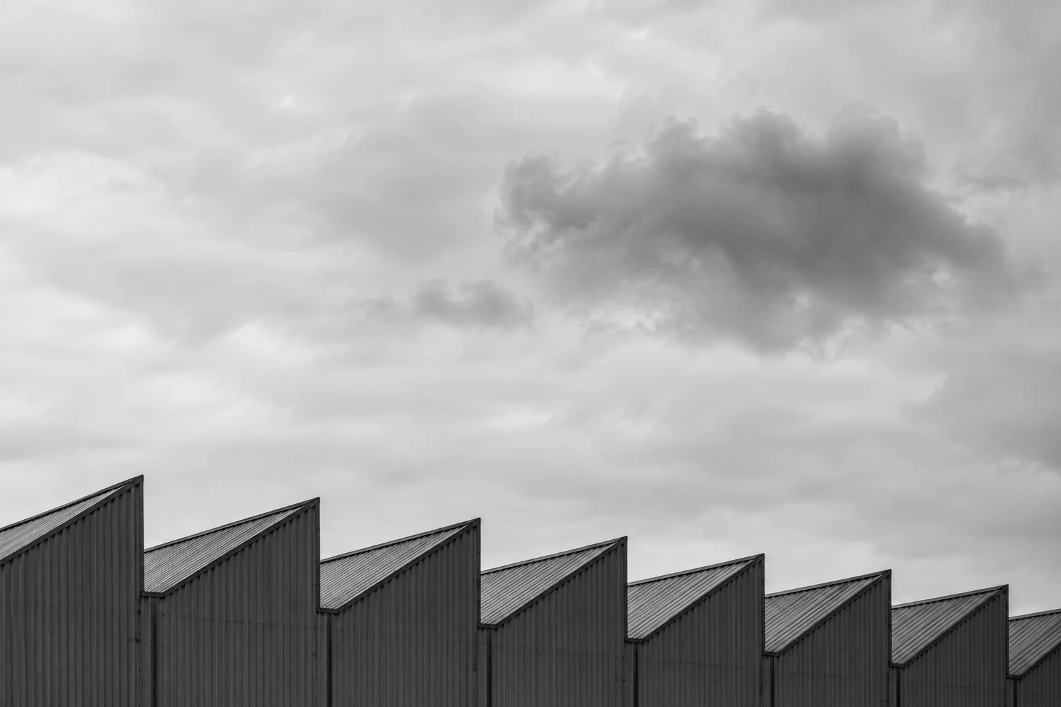 A receding row of corrugated industrial roof peaks against a pale overcast sky and one darker cloud.