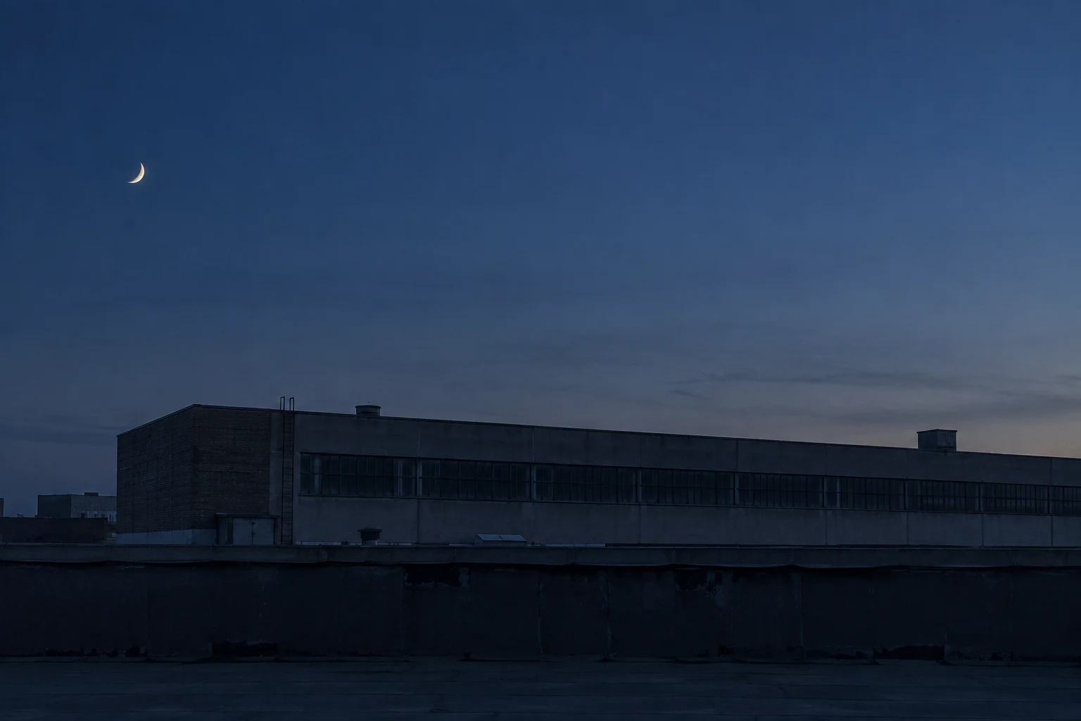 A small crescent moon in a deep blue sky above a long dark industrial roof and quiet parapet.