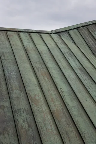 A close copper roof with green and brown weathering, standing seams and a shallow change of roof plane.