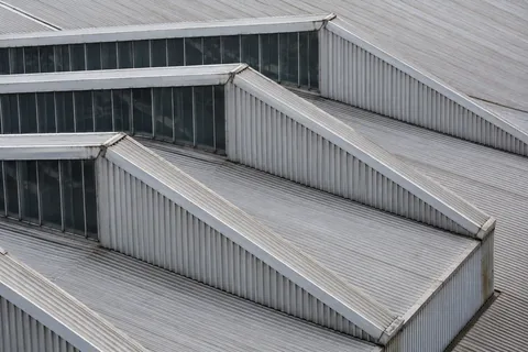 An elevated close view of industrial sawtooth roofs with corrugated metal slopes and narrow dark glazing.