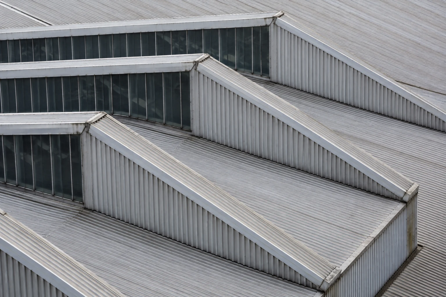 An elevated close view of industrial sawtooth roofs with corrugated metal slopes and narrow dark glazing.