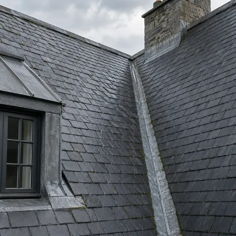 Two dark slate roof planes meeting at a metal-lined valley, beside a small dormer and stone chimney.