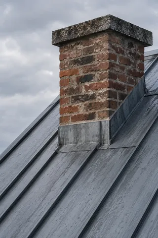 A short capped brick chimney stack with metal flashing on a gray standing-seam roof.