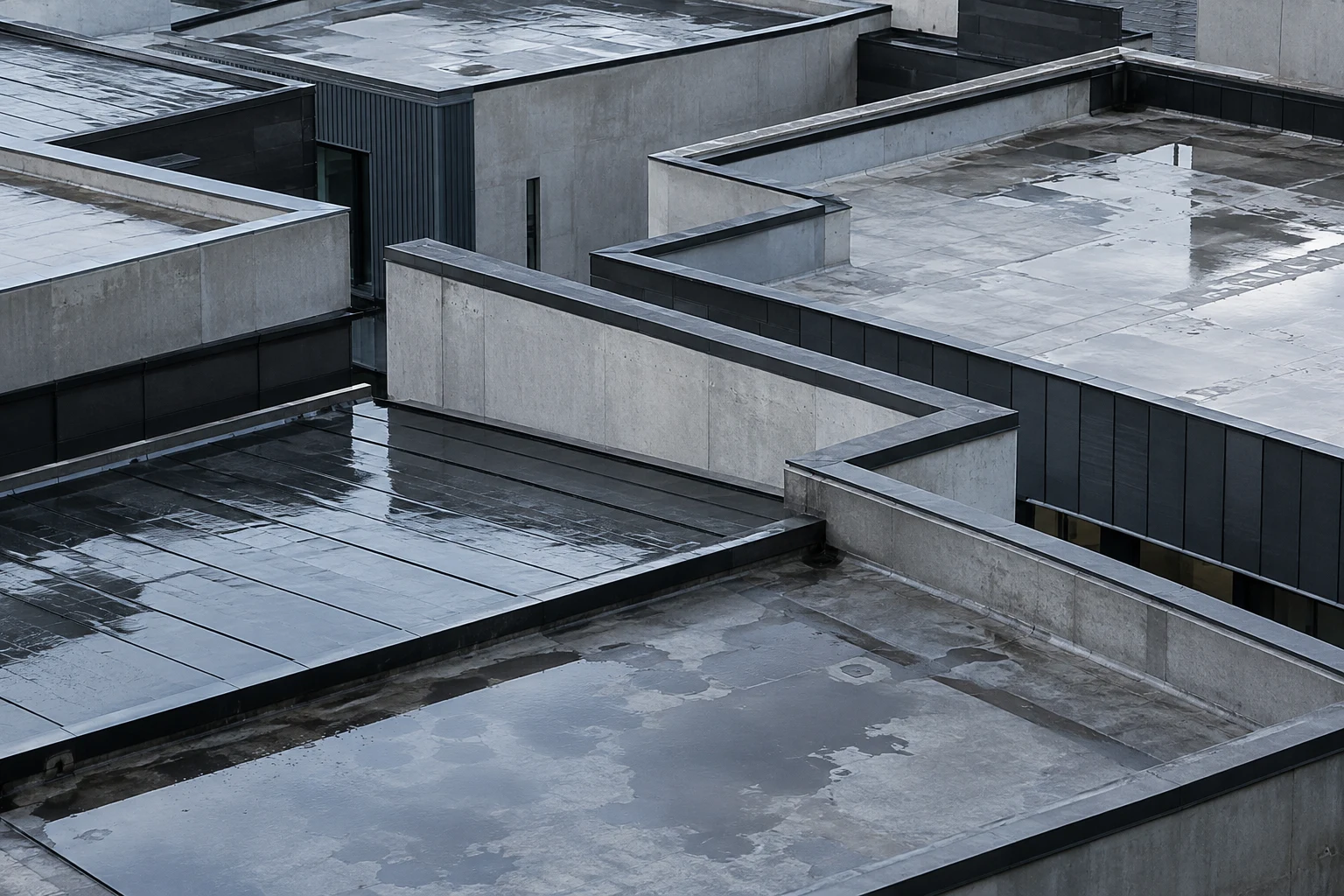 An elevated view of pale concrete and dark metal roof terraces with parapets and irregular shallow puddles.