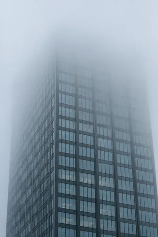 An anonymous blue-gray glass tower with aligned windows fading gradually into dense fog at the top.