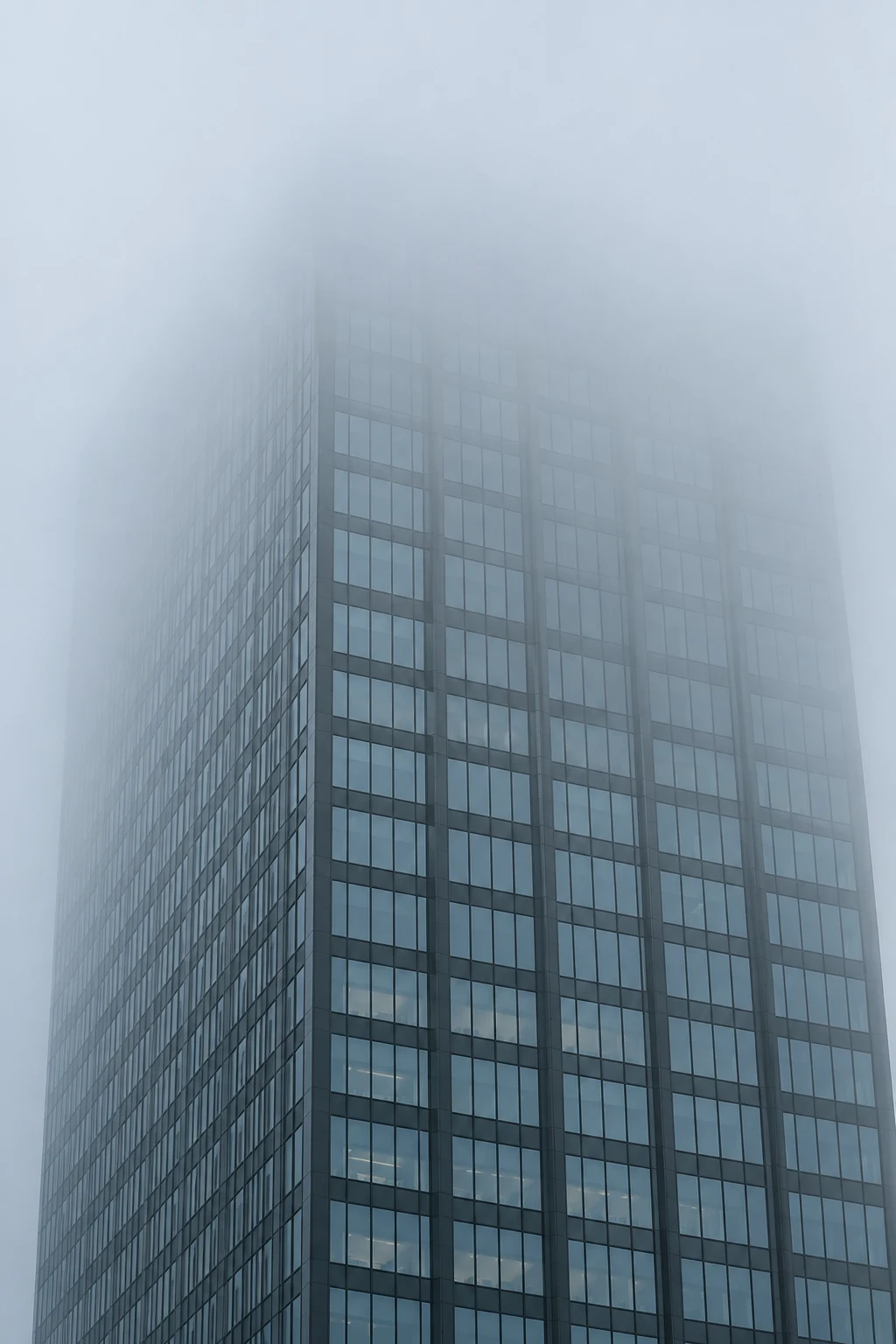 An anonymous blue-gray glass tower with aligned windows fading gradually into dense fog at the top.