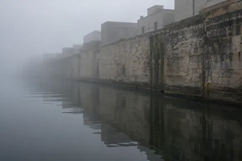 A long weathered stone canal wall with plain buildings above, reflected in calm dark water and disappearing into mist.