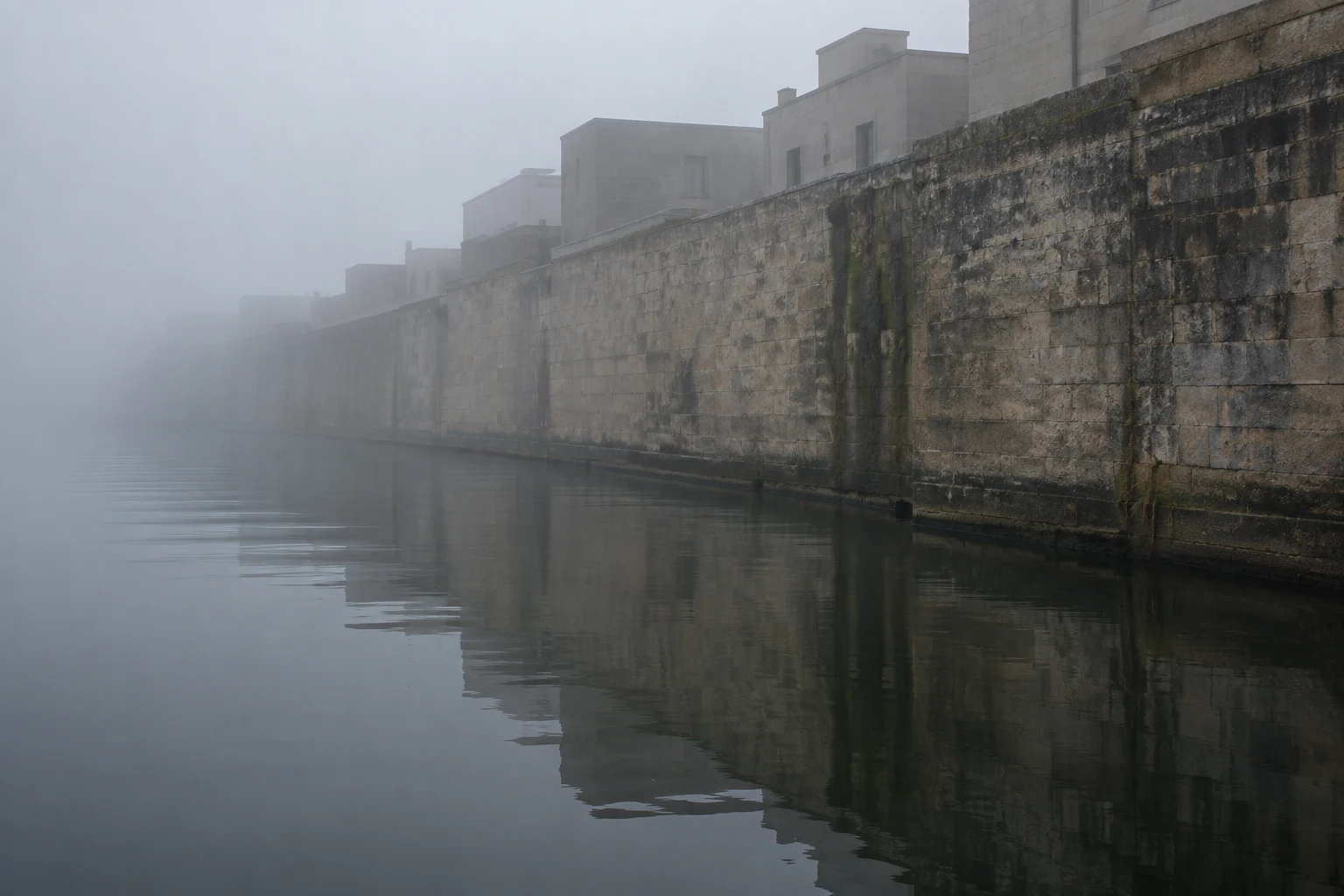 A long weathered stone canal wall with plain buildings above, reflected in calm dark water and disappearing into mist.