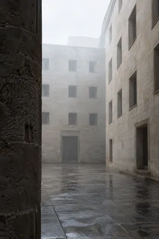 An empty misty courtyard with pale masonry buildings, square windows and wet paving, framed by a close dark wall.