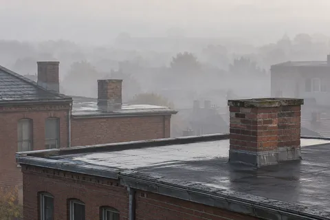 Three plain brick chimney stacks above damp urban rooftops with distant buildings and trees softened by fog.
