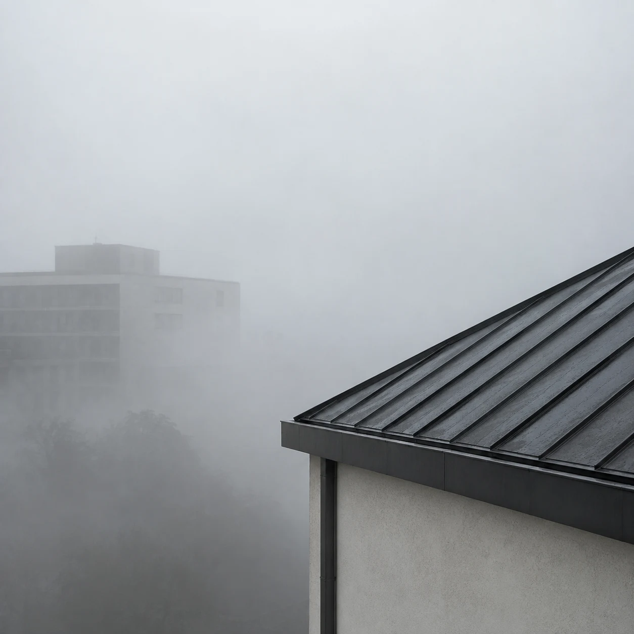 A sharp charcoal metal roof corner above a pale wall, with one faint block building in the fog beyond.