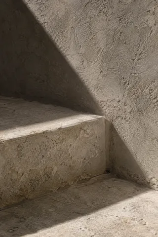 A broad diagonal shadow crosses a rough plaster wall and the corner of a pale stone step.