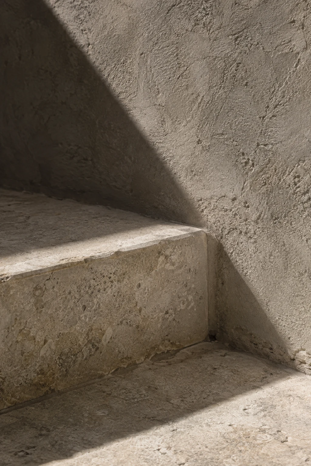 A broad diagonal shadow crosses a rough plaster wall and the corner of a pale stone step.